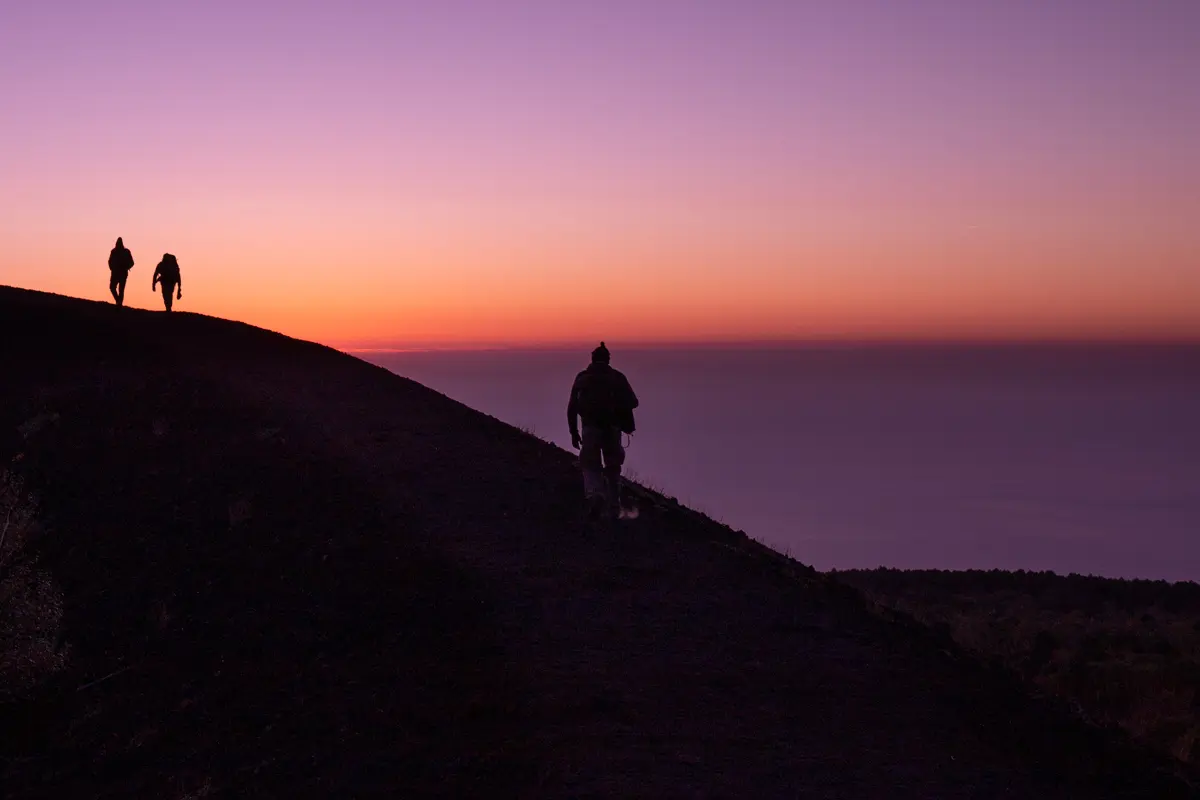 silhouette standing on a mountain with two other silhouettes far away on violet and orange sky monti sartorius at dawn mount etna