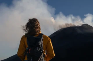 person standing in front of mount etna crater emitting smoke