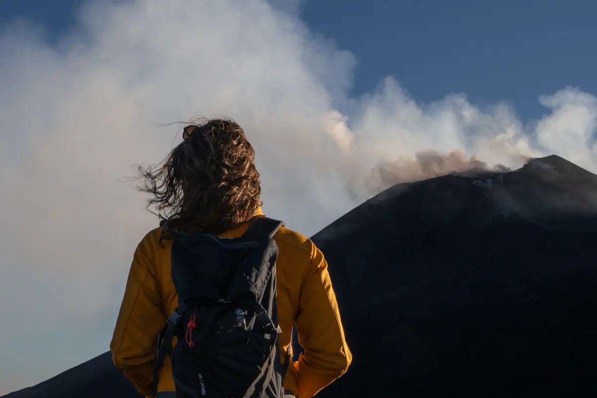 person standing in front of mount etna crater emitting smoke